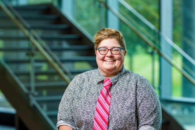 Dr. Amanda Jo Hobson has short cropped sandy blonde hair. She wears large-framed glasses, a black and white flower print shirt with a multi-hued pink tie. She smiles at the camera while standing in front of a flight of glass and steel stairs.
